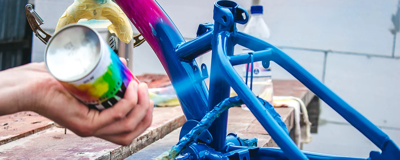 Farblack-Spraydose mit dem typischen gewölbten Spraydosen-Boden, mit der gerade ein Fahrradrahmen lackiert wird.